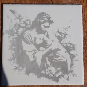 May include: Square ceramic tile featuring a grayscale image of a figure holding a lamb, surrounded by floral elements. The artwork has a vintage, classical style, with soft shading and detailed linework. The tile is set against a wooden surface.