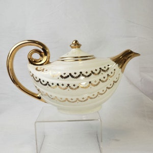 May include: White teapot with a gold handle and gold decorative details. The teapot has a rounded body and a spout. The teapot is sitting on a clear acrylic stand.
