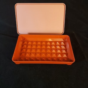 May include: A rectangular orange plastic container with a lid. The container has a textured bottom with a grid of small pyramids. The lid is a light pink color.