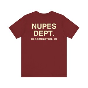 May include: A maroon t-shirt with the text "NUPES DEPT. BLOOMINGTON, IN" printed in a cream color. The shirt has short sleeves and a crew neck.