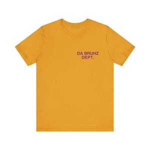 May include: Mustard yellow t-shirt with short sleeves. The text "DA BRUHZ DEPT." is printed in purple on the upper chest area. The shirt is made of a soft material and is suitable for casual wear.