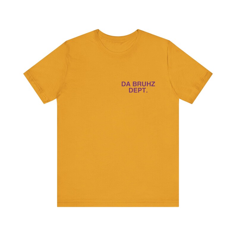 May include: Mustard yellow t-shirt with short sleeves. The text "DA BRUHZ DEPT." is printed in purple on the upper chest area. The shirt is made of a soft material and is suitable for casual wear.