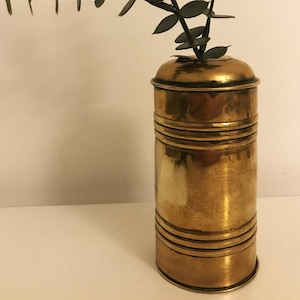 May include: A brass vase with a round, slightly flared top and a cylindrical body. The vase has a series of horizontal bands around the body. A sprig of eucalyptus with green leaves is in the vase.