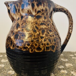 May include: A ceramic jug with a dark brown and black speckled pattern. The jug has a rounded body, a handle, and a spout. The bottom half is solid black, and the top half has a mottled design. It is likely used for serving beverages.