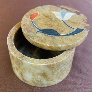 May include: A small, round, brown stone box with a lid. The lid features a floral design with black, white and red accents.