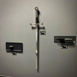 May include: A silver sword with a decorative hilt and a black handle is mounted on a gray wall. A black knife is mounted to the right. The sword has text etched on the blade. The items are displayed with small black magnetic holders.