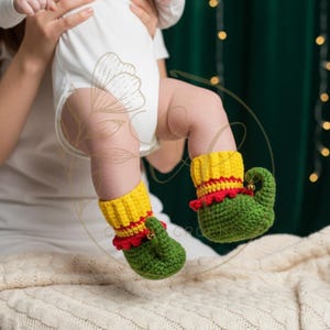 May include: A baby's legs are shown wearing handmade crochet elf booties. The booties are green with yellow cuffs and red trim. The baby is wearing a white onesie. The background is a blurred green with soft lighting.