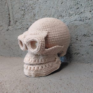 May include: A crocheted skull sculpture in a light beige color. The skull features detailed eye sockets, a prominent nose, and a defined jawline. The handmade piece is crafted with a textured stitch, giving it a unique, handcrafted appearance. This decorative item is suitable for Halloween or gothic decor.