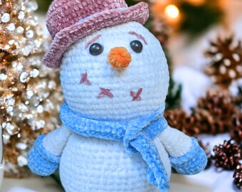 May include: A crocheted snowman toy with a pink velvet hat, blue velvet scarf, and blue mittens. The snowman is sitting on a white surface with a blurred background of a Christmas tree and pine cones.