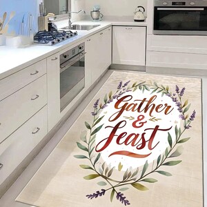 May include: A rectangular kitchen rug with the words "Gather & Feast" in a decorative script, surrounded by a watercolor wreath of green leaves and purple lavender sprigs. The rug has a light beige background.