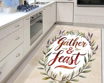 May include: A rectangular kitchen rug with the words "Gather & Feast" in a decorative script, surrounded by a watercolor wreath of green leaves and purple lavender sprigs. The rug has a light beige background.