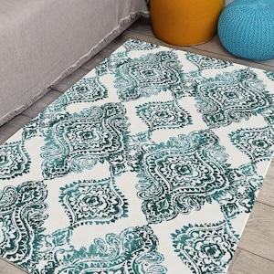 May include: A rectangular rug featuring a white base and a teal paisley design. The rug is positioned on a wooden floor, with a bed and decorative pouffes visible in the background. The pattern is a repeating design of stylized floral and leaf motifs.