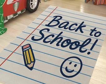 May include: A rectangular rug with a notebook paper design. The rug features blue horizontal lines, a red vertical line, and the words "Back to School!" in dark blue. A yellow pencil and a smiley face are also on the rug.