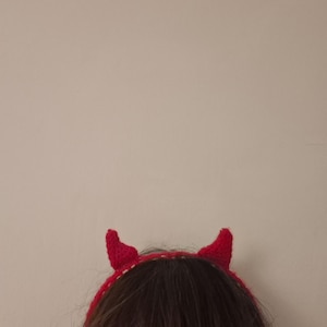 May include: A bright red crocheted headband featuring two small, pointed devil-style horns. The accessory is constructed from yarn with a visible textured stitch pattern, designed to sit atop the head with the horns protruding upwards.