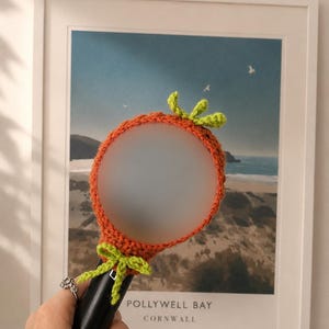 May include: A circular hand mirror featuring a crocheted orange cover with a green leaf accent at the top and a green bow at the base. The mirror surface is frosted, and the handle is black. The design resembles a carrot, providing a textured finish.