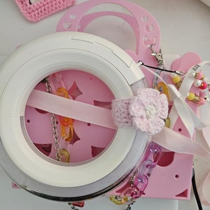 May include: A white ring light with a silver trim and a pink crocheted flower attached to it. The ring light is on a pink surface with a white ribbon and a chain of colorful plastic links.