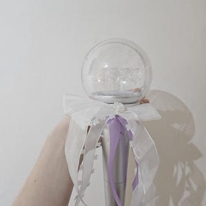 Pode incluir: Uma varinha luminosa branca e roxa com uma esfera de plástico transparente no topo. A esfera é cheia de glitter e tem um laço branco amarrado ao redor.