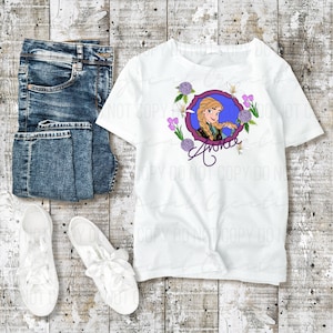 May include: White t-shirt featuring a cartoon image of a character with blonde braids, surrounded by flowers, and the name "Anna". The shirt is paired with blue jeans and white sneakers on a wooden surface.