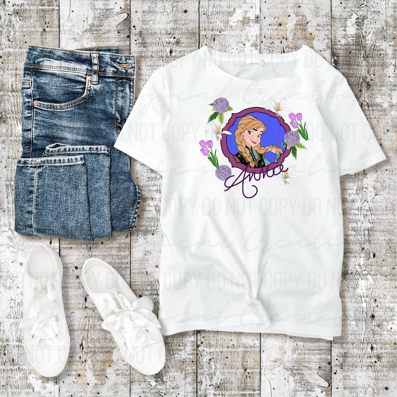 May include: White t-shirt featuring a cartoon image of a character with blonde braids, surrounded by flowers, and the name "Anna". The shirt is paired with blue jeans and white sneakers on a wooden surface.