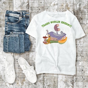 May include: White t-shirt with the text "TRASH GOBLIN ENERGY!" in green. The shirt features a cartoon illustration of a purple creature lounging on a pile of discarded food, including an apple core and banana peel. Blue jeans and white sneakers complete the casual outfit.