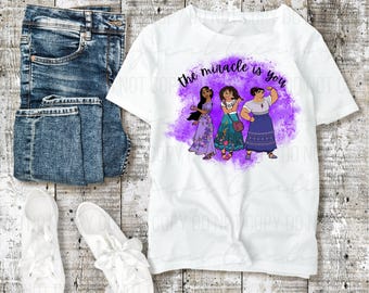 May include: White t-shirt with the text "the miracle is you" and a graphic of three cartoon women in colorful dresses. The shirt is paired with blue jeans and white sneakers on a wooden surface.