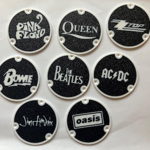 May include: Eight round black and white stickers with the names of famous bands: Pink Floyd, Queen, ZZ Top, Bowie, The Beatles, AC/DC, Jimi Hendrix, and Oasis.