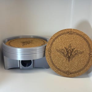May include: A silver-colored engine piston-shaped holder containing round cork coasters. Each coaster features a laser-engraved design of a stylized V8 emblem surrounded by a circular border with pointed accents at the top and bottom edges.