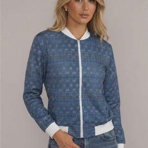 Peut inclure: Un blouson bleu orné d'un motif répétitif de cercles dorés entrelacés sur tout le tissu. La veste présente une fermeture éclair blanche sur le devant, un col, des poignets et un ourlet à la taille en bord-côte de couleur blanche.