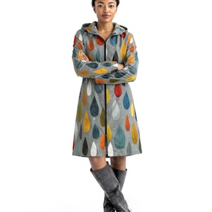 Puede incluir: Un impermeable con capucha hasta la rodilla en color gris claro con un patrón repetitivo de gotas en tonos amarillo mostaza, naranja quemado, azul oscuro y blanco, con una cremallera frontal negra y botas altas de color gris oscuro.