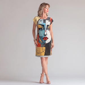 May include: A knee-length shift dress featuring a bold, abstract face graphic in shades of blue, yellow, red, and black. The design includes a stylized eye, red lips, and geometric lines printed across the front of the garment, which has short sleeves and a round neckline.