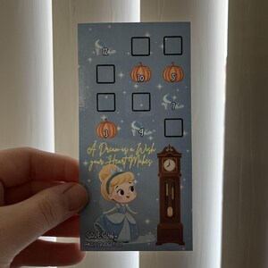 May include: A light blue card featuring Cinderella, pumpkins, and glass slippers. The card has a clock and the text "A Dream is a Wish Your Heart Makes." It includes numbers and blank squares for a game or activity.
