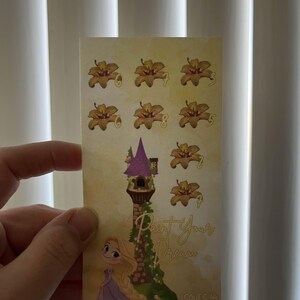 May include: A rectangular sticker featuring a cartoon illustration of a tower with a purple roof and a blonde girl in a purple dress. The sticker has the text "Paint Your Dream" and several flower illustrations.
