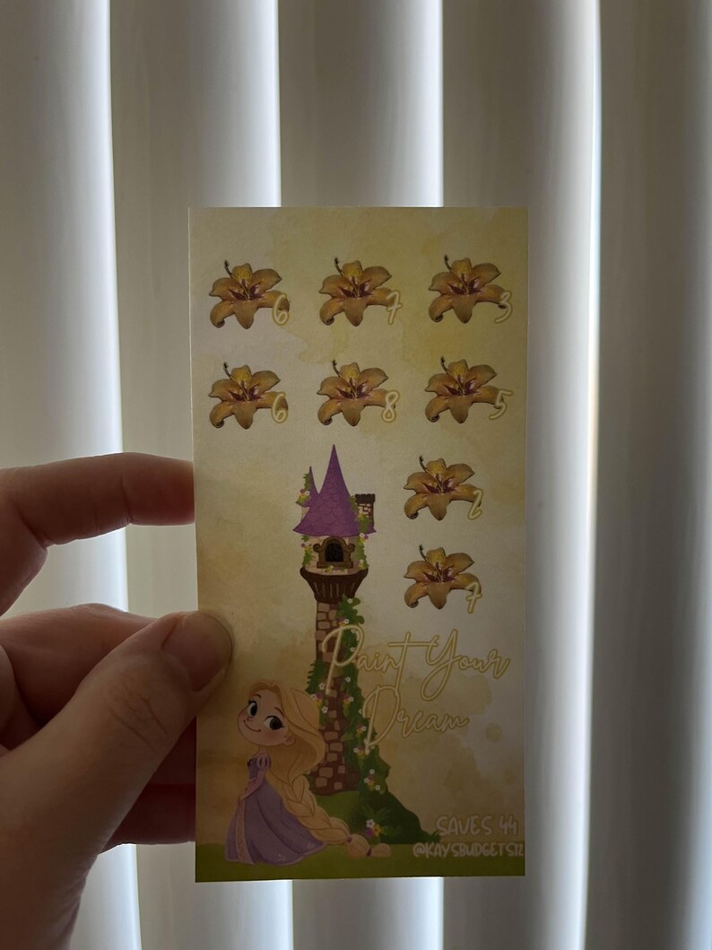 May include: A rectangular sticker featuring a cartoon illustration of a tower with a purple roof and a blonde girl in a purple dress. The sticker has the text "Paint Your Dream" and several flower illustrations.