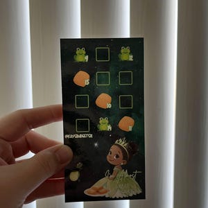 May include: A rectangular card with a dark green background featuring a cartoon illustration of a princess in a green dress. The card has a grid of squares with frog and food illustrations, and the text "I'm Almost There" and "@kaysbudgetsz".
