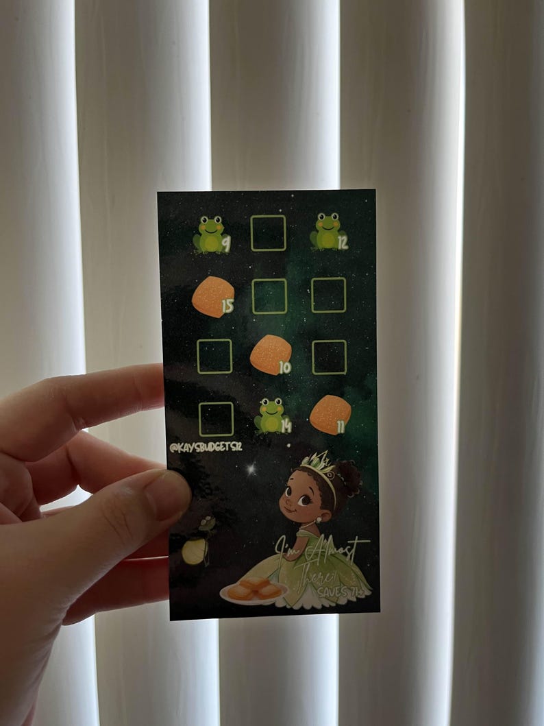 May include: A rectangular card with a dark green background featuring a cartoon illustration of a princess in a green dress. The card has a grid of squares with frog and food illustrations, and the text "I'm Almost There" and "@kaysbudgetsz".