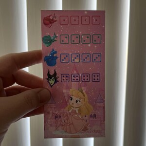 May include: A pink card featuring illustrations of characters from a fairy tale. The card has dice-like graphics and the text "Once Upon a Dream". The characters include a princess and fairies.