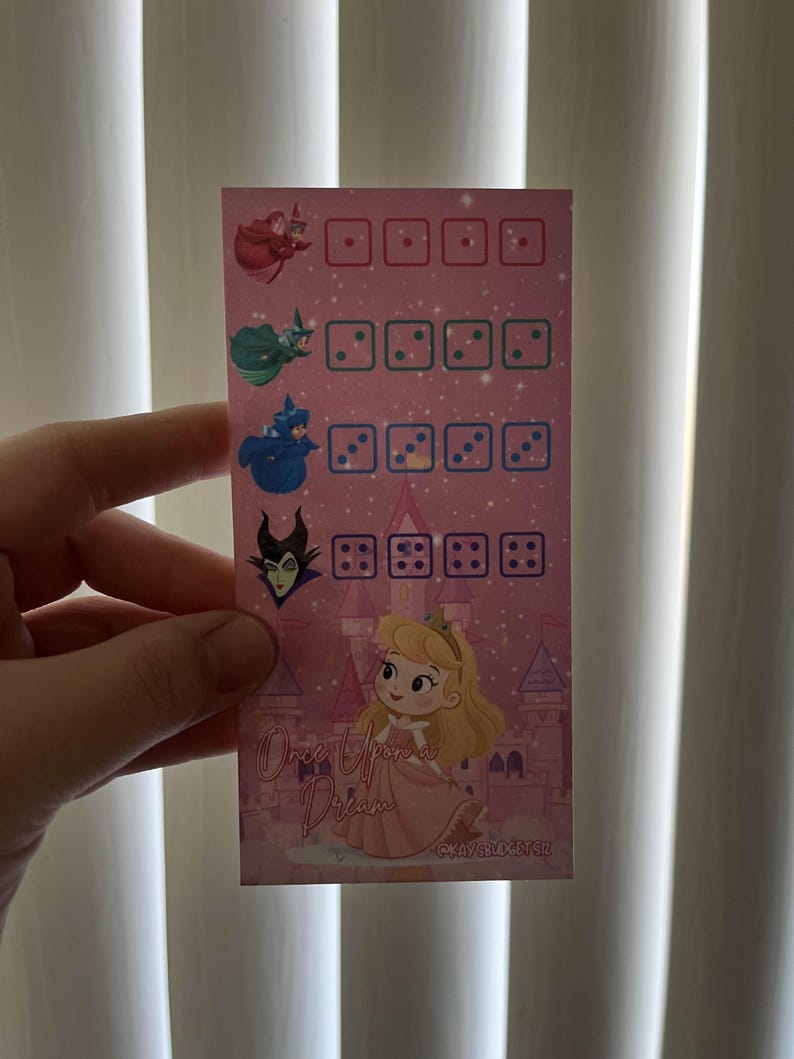 May include: A pink card featuring illustrations of characters from a fairy tale. The card has dice-like graphics and the text "Once Upon a Dream". The characters include a princess and fairies.