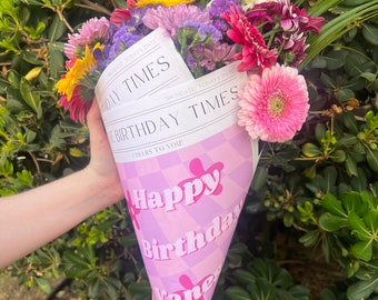 May include: A pink and purple cone-shaped bouquet wrapper with the text "Happy Birthday" and "Cheers to You!"  The wrapper is decorated with a checkered pattern and a pink flower. A bouquet of colorful flowers is inside the wrapper.