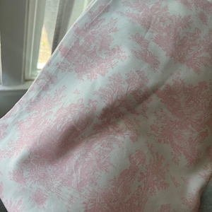 May include: A folded piece of fabric featuring a pink and white toile pattern. The design depicts pastoral scenes with figures, trees, and architectural elements. The fabric has a soft, slightly textured appearance.