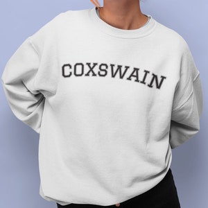 Puede incluir: Sudadera blanca con la palabra "COXSWAIN" impresa en negro, arqueada sobre el pecho. La sudadera tiene cuello redondo y mangas largas. El fondo es de color azul claro liso.