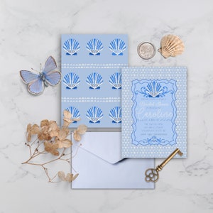 Puede incluir: Una invitación azul y blanca con estampado de conchas marinas para una despedida de soltera para Caroline. La invitación incluye la fecha, la hora y el lugar del evento. La invitación está rodeada de un sobre azul, una llave, una concha y una mariposa.