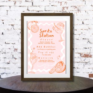 Peut inclure: Une affiche rose encadr&eacute;e avec des illustrations et du texte orange. L'affiche indique "Spritz Station" avec des instructions pour faire un cocktail spritz, y compris les ingr&eacute;dients et les garnitures. Le cadre est de couleur marron fonc&eacute;.