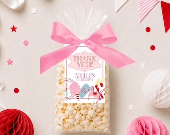 May include: A clear cellophane bag filled with popcorn, tied with a pink ribbon. A decorative tag reads "Thank You!" and "Adelle's 5th Birthday" with carnival-themed illustrations. Pink and red confetti and decorations surround the bag.