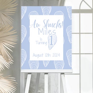 May include: A light blue birthday sign with a white oyster shell pattern. The sign says "Aw Shucks! Miles is turning 1 August 12th 2024".