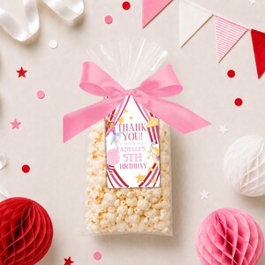 May include: A clear cellophane bag filled with popcorn, tied with a pink ribbon and a tag that reads "THANK YOU!" and "Adelle's 5th Birthday". The tag has a circus theme with red and white stripes and star accents.