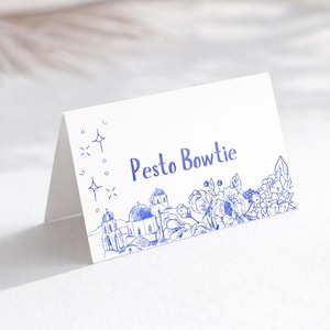 May include: A folded white place card with blue ink illustrations. The card features the text "Pesto Bowtie", a floral design, and a cityscape with domes and towers. Decorative starbursts are on the left.
