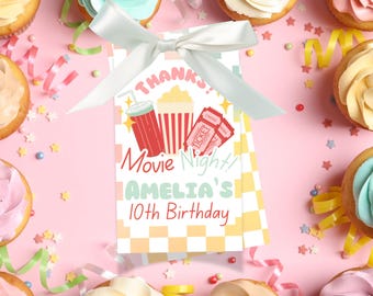 Op de afbeelding: Een feestelijke afbeelding met cupcakes en een cadeaulabel vastgebonden met een wit lint. Het label leest "Bedankt! Movie Night! Amelia's 10e verjaardag" met illustraties van popcorn, een drankje en bioscoopkaartjes. De achtergrond is roze.