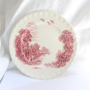 May include: A white ceramic dinner plate with a pink floral design featuring a cottage by a lake with birds flying overhead.