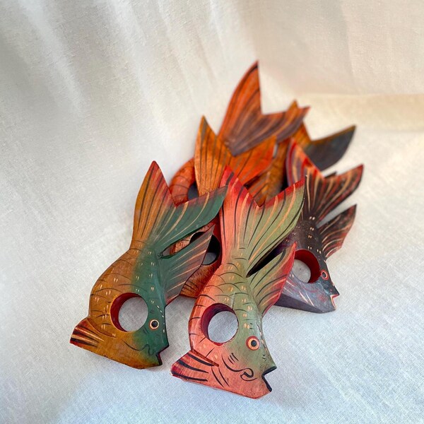 Puede incluir: Un conjunto de servilleteros de madera con forma de pez. Cada servilletero está pintado a mano con una combinación de colores única, con tonos de verde, naranja y rojo. Los peces tienen un agujero para la servilleta y están colocados sobre una superficie blanca.