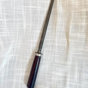 May include: A steel knife sharpening rod with a dark wooden handle and a silver-colored metal ring. The rod is tapered and appears well-used. The handle is a deep, rich brown color.